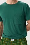 A man wearing a dark green short-sleeve crewneck t-shirt with a chest pocket, paired with green and yellow plaid pajama pants. The brand label on the pants reads 'Bambi & Brooke'.