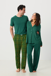 A man and a woman modeling matching green pajamas against a plain white studio background. The man wears a short-sleeve green shirt and green plaid pajama pants, while the woman wears a green button-front pajama top and matching green pajama pants.