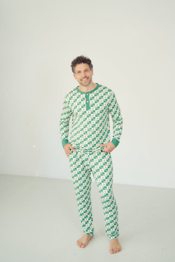 A man wearing a green and white checkered long-sleeve pajama set with a green Henley collar, standing barefoot against a plain white studio background.