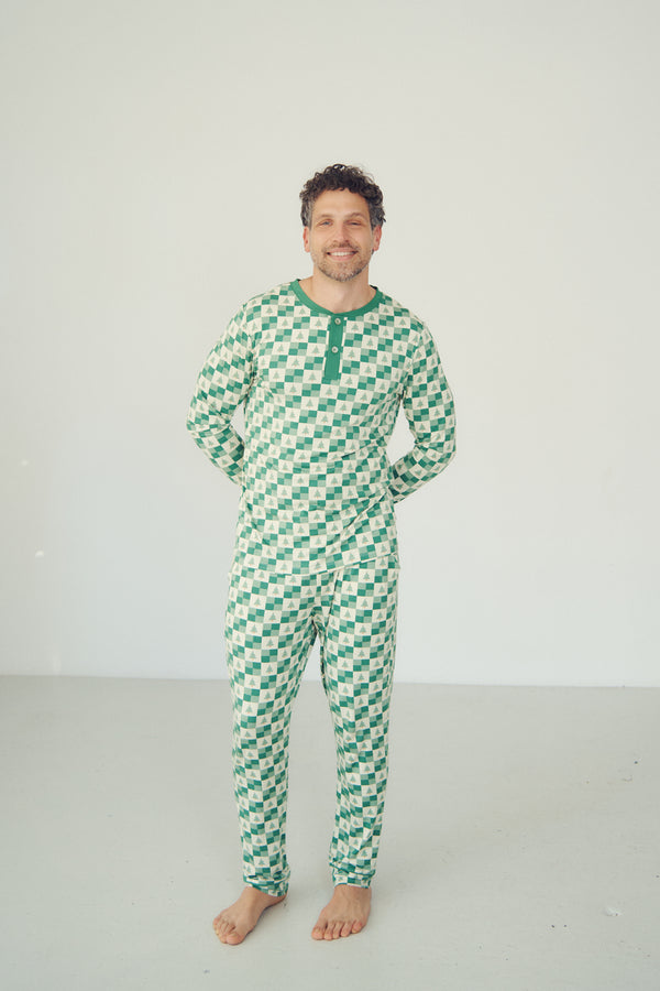 A man wearing a green and white patterned long-sleeve pajama top and matching pants, standing barefoot against a plain light gray studio background.