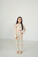 A young girl stands barefoot in a bright, minimalist studio setting, wearing a cream-colored long-sleeve pajama set with a festive Christmas print of red ornaments, green pine trees, and white snowflakes.