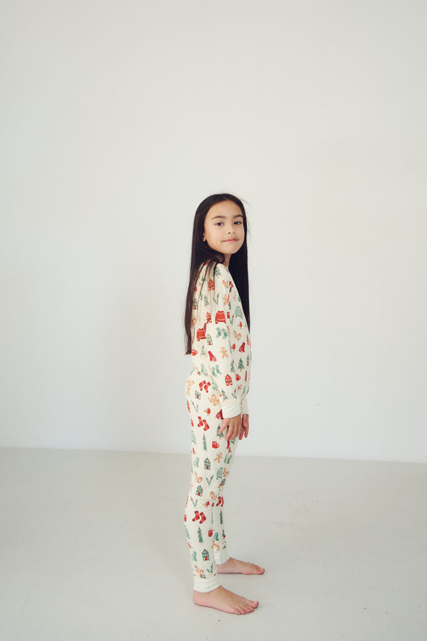 A young girl stands barefoot in profile wearing a long-sleeve pajama set with a festive holiday print of red ornaments, green trees, and white snowflakes on an off-white background, set against a plain white studio wall.
