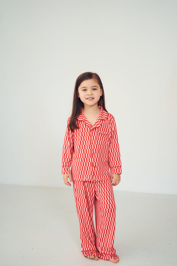 A young girl with long dark hair stands barefoot against a plain white studio background, wearing a matching red and white striped pajama set with a long-sleeve top and pants.