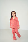 A young girl with long dark hair stands barefoot in a red and white patterned pajama set against a plain light gray studio background.