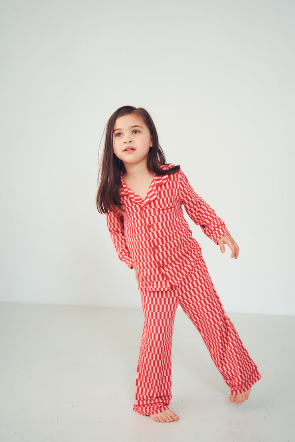A young girl with long brown hair stands barefoot in a studio setting, wearing a red and white patterned long-sleeve pajama set with a matching top and pants.