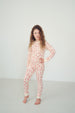 A young girl with long curly brown hair stands barefoot in a studio setting, wearing a long-sleeve one-piece pajama set with an allover red and cream pattern.
