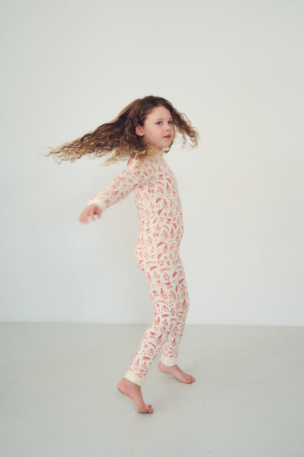 A young girl with long, curly brown hair is captured mid-motion while wearing a cream-colored one-piece pajama or bodysuit with a red abstract pattern, set against a plain light gray studio background.