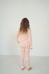 A woman with long curly brown hair stands with her back to the camera wearing a matching two-piece pajama set with a pink and white floral pattern, set against a plain white studio background.
