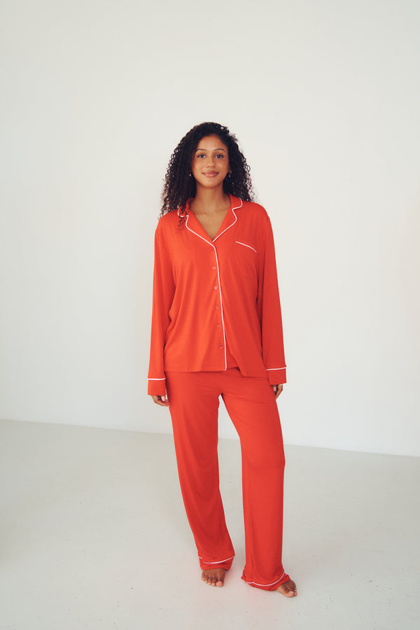 A woman modeling a bright orange-red pajama set with white piping details, consisting of a long-sleeve button-up top and matching pants, set against a plain white studio background.