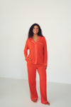 A woman modeling a bright orange-red pajama set with white piping details, standing against a plain white studio background.