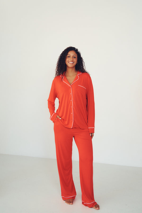 A woman modeling a bright orange-red pajama set with white piping details, standing against a plain white studio background.