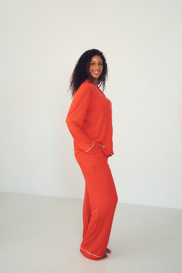 A woman modeling a bright orange-red two-piece pajama set with white piping, standing in profile against a clean white studio background.