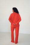 A woman viewed from behind wearing a bright orange-red pajama set with white piping trim, standing against a plain white studio background.