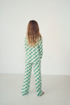 A person viewed from behind wearing a green and white checkered long-sleeve jumpsuit or pajama set, standing against a plain white wall in a minimalist indoor setting.