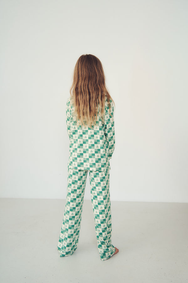 A person viewed from behind wearing a green and white checkered long-sleeve jumpsuit or pajama set, standing against a plain white wall in a minimalist indoor setting.