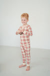 A young boy with red hair stands barefoot in a studio wearing a long-sleeve plaid pajama top and matching pants in cream, red, and green.