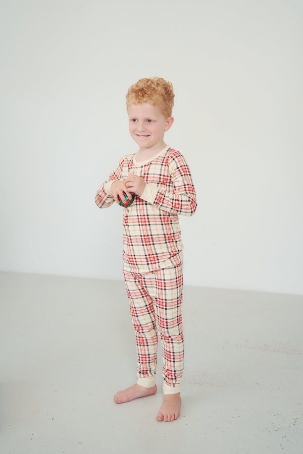 A young boy with red hair stands barefoot in a studio wearing a long-sleeve plaid pajama top and matching pants in cream, red, and green.