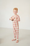 A young boy with red hair stands barefoot in a studio wearing a long-sleeve plaid pajama set in cream with red and orange lines.