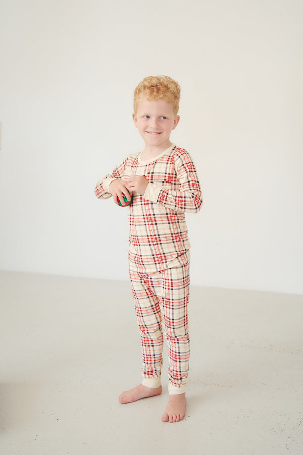 A young boy with red hair stands barefoot in a studio wearing a long-sleeve plaid pajama set in cream with red and orange lines.