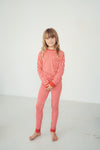 A young girl with blonde hair stands barefoot in a white studio setting, wearing a red and white patterned long-sleeve pajama set with solid red cuffs and collar.
