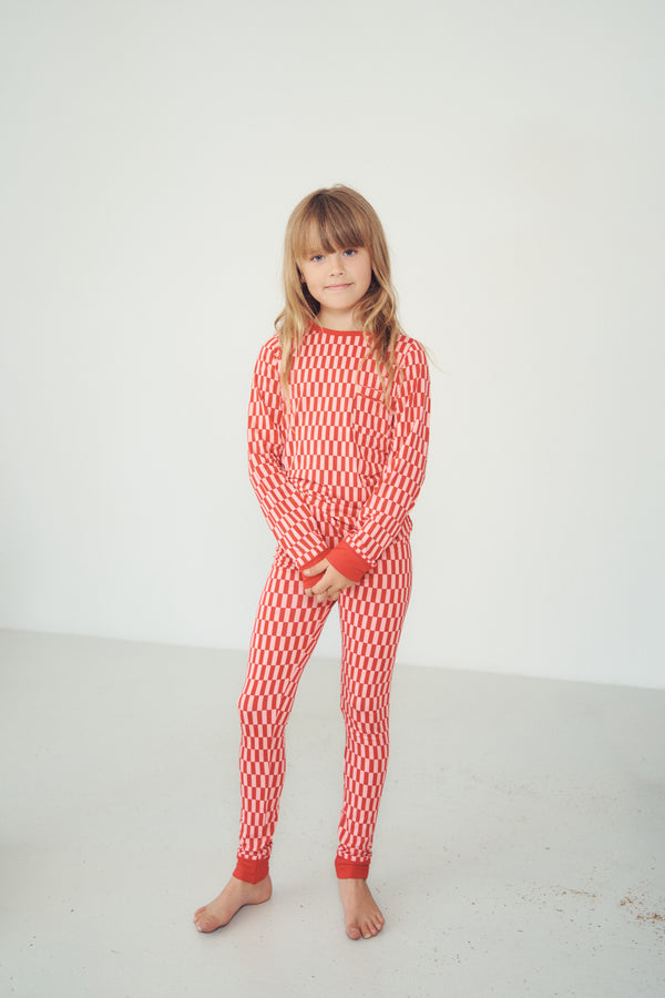 A young girl with blonde hair stands barefoot in a white studio setting, wearing a red and white patterned long-sleeve pajama set with solid red cuffs and collar.