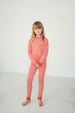 A young girl with blonde hair stands barefoot in a white studio setting, wearing a red and white patterned long-sleeve pajama set with solid red cuffs and collar.