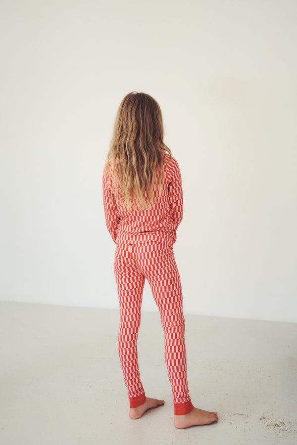 A child standing with their back to the camera, wearing a matching red and white patterned long-sleeve top and pants set in a minimalist studio setting.