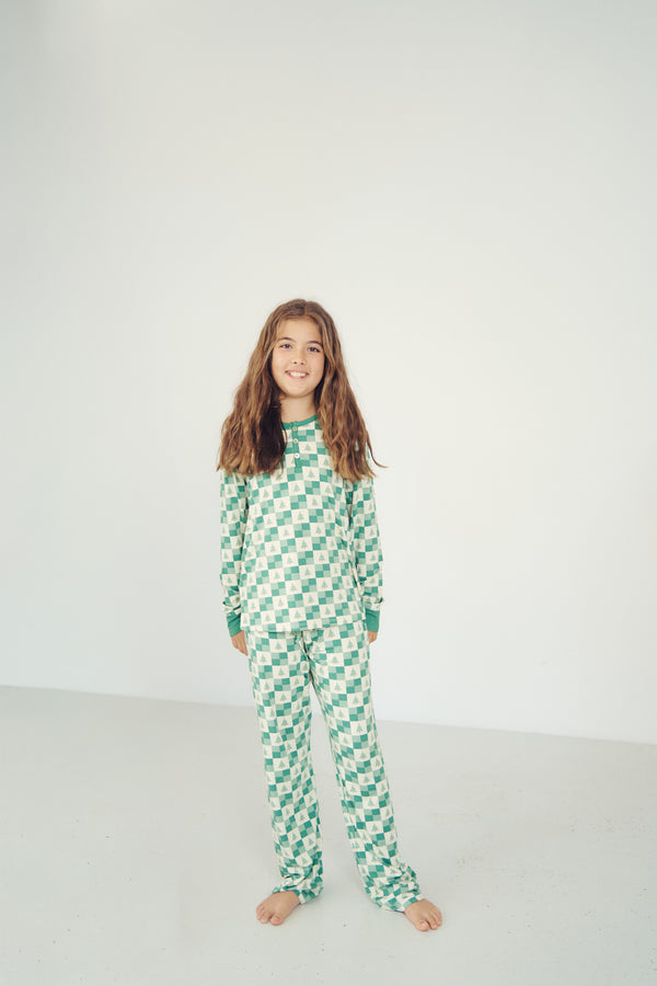 A young girl with long brown hair stands barefoot in a bright white studio, wearing a green and white checkered long-sleeve pajama set.
