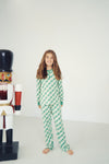 A young girl with long light brown hair stands barefoot in a white studio, wearing a green and white checkered long-sleeve pajama set. To her left is a large nutcracker figurine.