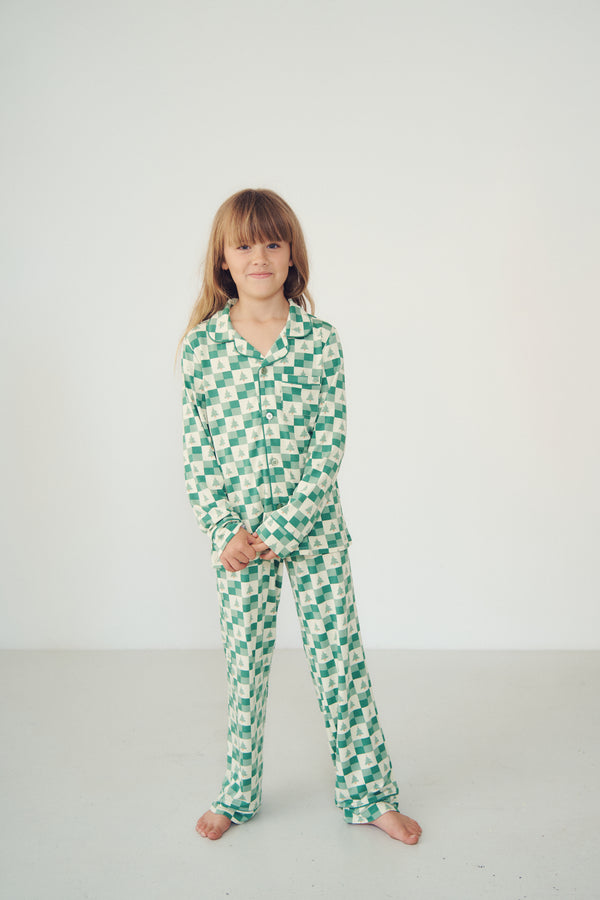 A young girl with light brown hair stands barefoot in a studio setting, wearing a matching green and white patterned pajama set consisting of a long-sleeve top and pants.