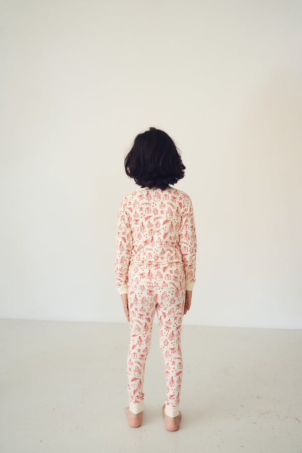 A child viewed from behind wearing a long-sleeve one-piece jumpsuit with a dense coral-red floral pattern on a cream base, standing against a plain off-white wall.