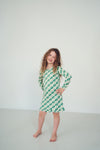A young girl with curly brown hair stands barefoot in a studio, wearing a green and white checkered long-sleeve dress with a ruffled hem against a plain light gray background.