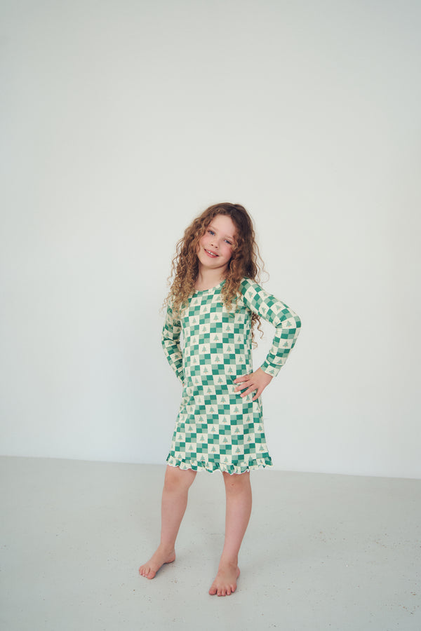 A young girl with curly brown hair stands barefoot in a studio, wearing a green and white checkered long-sleeve dress with a ruffled hem against a plain light gray background.