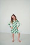 A young girl with curly brown hair stands barefoot in a studio, wearing a green and white checkered long-sleeve dress with a boat neckline.