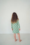 A child with long curly brown hair stands barefoot from the back, wearing a green and white checkered long-sleeve dress against a plain white wall and light gray floor.