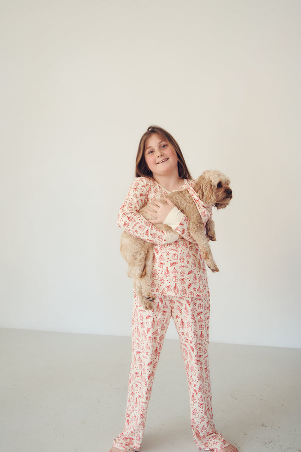 A young girl with long brown hair stands in a bright studio, wearing a light pink and white patterned pajama set and holding a small fluffy tan dog.