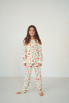 A young girl with long brown hair stands barefoot against a plain white studio background, wearing a long-sleeve pajama top and matching pants with a small repeating holiday-style print in red, green, and blue on an off-white base.