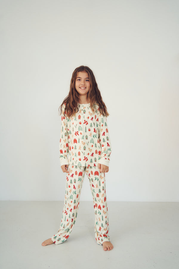 A young girl with long brown hair stands barefoot against a plain white studio background, wearing a long-sleeve pajama top and matching pants with a small repeating holiday-style print in red, green, and blue on an off-white base.