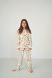 A young girl with long brown hair stands barefoot against a plain white studio background, wearing a long-sleeve pajama top and matching pants with a small repeating holiday-style print in red, green, and blue on an off-white base.