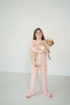 A young girl with long brown hair stands barefoot in a white studio, wearing a matching long-sleeve pajama set with a red abstract pattern. She is holding a small fluffy tan dog.