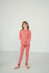A young girl with light brown hair stands barefoot in a studio setting, wearing a red and white patterned long-sleeve pajama set against a plain white background.