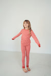 A young girl with light brown hair stands barefoot in a studio setting, wearing a matching red and white patterned long-sleeve top and pants set.