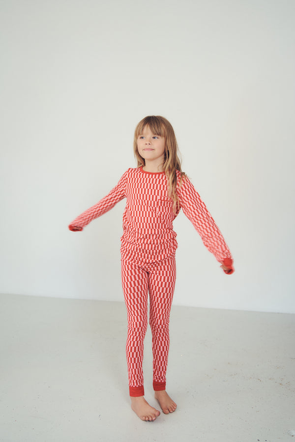 A young girl with light brown hair stands barefoot in a studio setting, wearing a matching red and white patterned long-sleeve top and pants set.