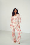 A woman modeling a two-piece plaid pajama set consisting of a long-sleeve button-up top and matching pants, in a red, orange, and white plaid pattern against a plain white studio background.