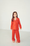 A young girl stands barefoot in a bright studio wearing a matching red pajama set with white piping trim. The set includes a long-sleeve button-front top with a chest pocket and matching wide-leg pants.