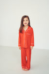 A young girl with long brown hair stands barefoot in a bright studio setting, wearing a matching red-orange pajama set with white piping details.