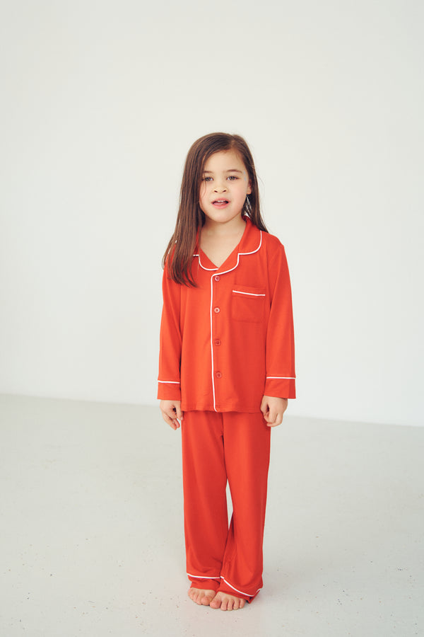 A young girl with long brown hair stands barefoot in a bright studio setting, wearing a matching red-orange pajama set with white piping details.