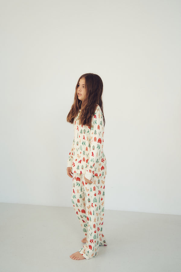 A young girl stands in profile wearing a cream-colored pajama set with a festive red and green Christmas tree pattern, set against a plain white studio background.