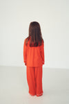 A child standing with their back to the camera, wearing a bright orange-red pajama set with white piping trim on a plain white studio background.