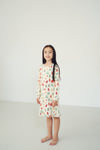A young girl with long dark hair stands barefoot in a minimalist white studio setting, wearing a long-sleeve cream-colored nightgown decorated with a colorful Christmas tree pattern.